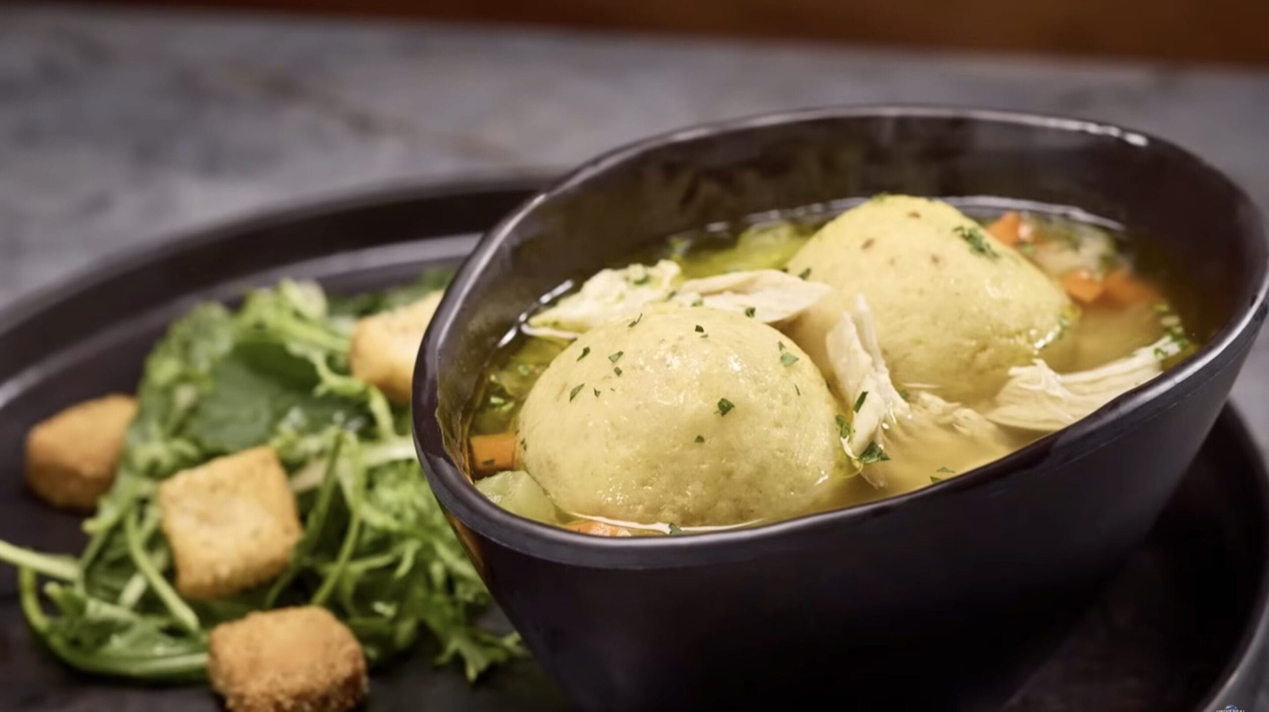 A bowl of matzo ball soup with vegetables and chicken sits on a plate, paired with a mixed green salad topped with croutons. This comforting dish is a delightful part of the Epic Universe of food and beverage.